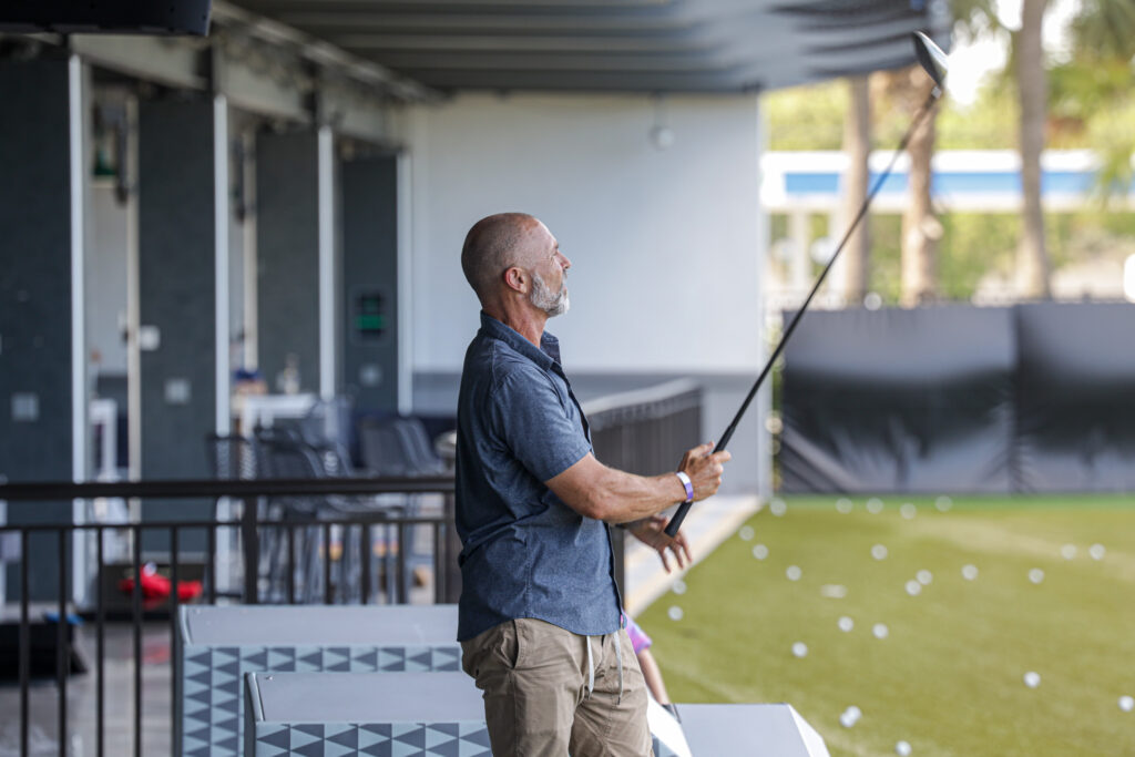 A man at drive shack holds a golf club on the bay, watching where his ball landed.